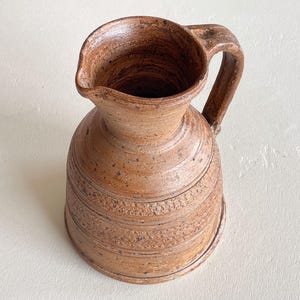 Puede incluir: Jarra artesanal de cerámica marrón con asa. La jarra tiene una base redondeada y un cuello estrecho con pico. Líneas horizontales decorativas rodean el cuerpo. La jarra mide aproximadamente 18 cm de altura.