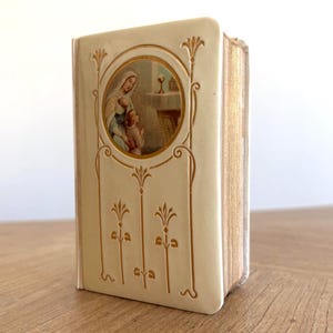 Peut inclure: Un livre couleur crème avec des détails floraux dorés en relief et une illustration circulaire encastrée sur la couverture. L'image montre une silhouette voilée et un enfant en prière devant un autel surmonté d'un calice.
