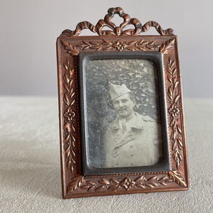 Peut inclure: Un cadre photo vintage en métal cuivré contenant une photographie en noir et blanc. Le cadre présente un design orné de détails floraux et de feuilles, ainsi qu'un élément décoratif en forme de ruban en haut. La photo montre un homme en uniforme militaire.