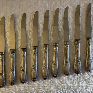 May include: Eight silver dinner knives with ornate handles. The knives have a polished finish and are arranged in a row on a white textured surface.