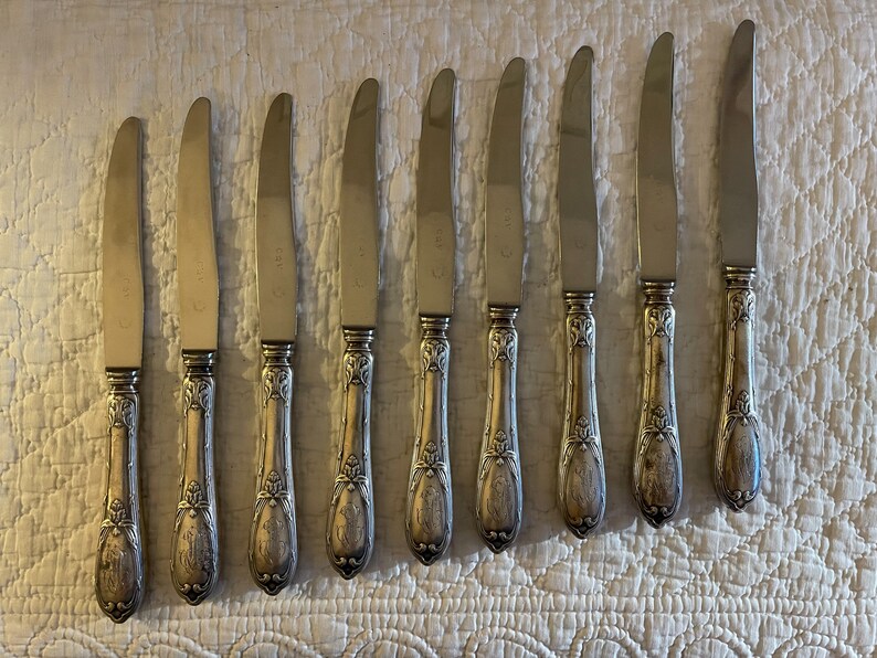 May include: Eight silver dinner knives with ornate handles. The knives have a polished finish and are arranged in a row on a white textured surface.