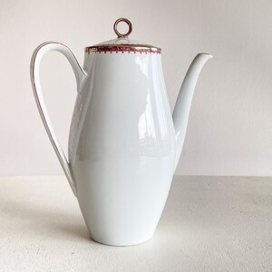 Puede incluir: Cafetera de cerámica blanca con asa y boquilla curvadas. La tapa tiene un asa de anillo dorado y un ribete rojo y dorado. El asa y el ribete tienen detalles dorados. La cafetera es de diseño clásico.