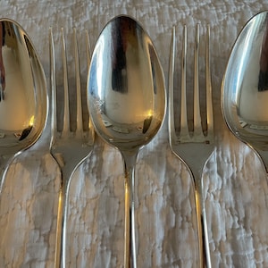 Peut inclure: Un ensemble de cuillères et de fourchettes en argent, disposées en rangée sur une surface texturée blanche. Les cuillères et les fourchettes sont polies et réfléchissent la lumière.