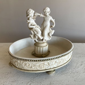 Può includere: Piatto decorativo in ceramica bianca con una statuetta scolpita di due bambini che ballano in cima. Il piatto ha un bordo ondulato e un design in rilievo attorno al bordo.
