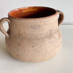 Puede incluir: Una olla de cerámica beige con dos asas curvas. El interior es de color marrón rojizo. La olla tiene un cuerpo redondeado y una abertura ancha. El exterior tiene una textura ligeramente rugosa y algunas marcas. La olla tiene un sello que dice "VILLASIS, S.A.M."