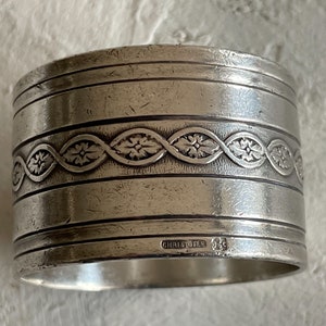May include: A cylindrical silver napkin ring featuring a central band with an embossed floral vine pattern. The surface has horizontal engraved lines and a visible hallmark stamp that reads CHRISTOFLE along with a small square emblem.