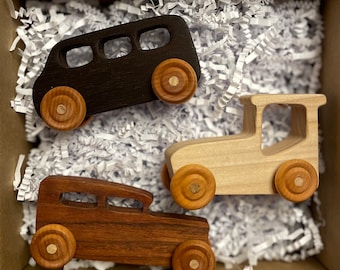 May include: Three wooden toy cars in a cardboard box with white paper filler. The cars are a black van, a light-colored truck, and a brown vintage car. All have wooden wheels.