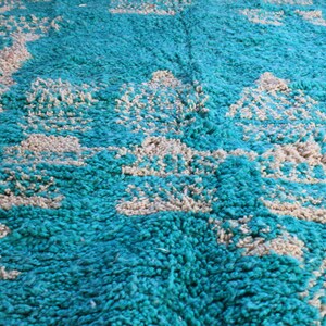 May include: Close-up of a teal and beige rug. The rug has a shaggy texture with a pattern of beige shapes against a teal background. The rug appears to be made of a soft, plush material, suitable for a living room or bedroom.