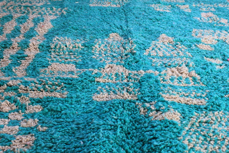 May include: Close-up of a teal and beige rug. The rug has a shaggy texture with a pattern of beige shapes against a teal background. The rug appears to be made of a soft, plush material, suitable for a living room or bedroom.