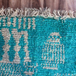 May include: A rectangular rug with a deep teal shag texture featuring cream-coloured geometric patterns and symbols. The top edge is finished with a row of cream-coloured fringe tassels, creating a textured surface with a high-pile appearance.