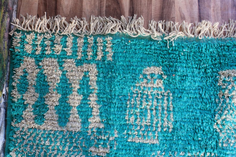 May include: A rectangular rug with a deep teal shag texture featuring cream-coloured geometric patterns and symbols. The top edge is finished with a row of cream-coloured fringe tassels, creating a textured surface with a high-pile appearance.