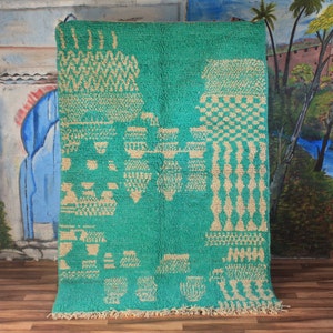 May include: A turquoise rug with beige geometric patterns. The rug has various shapes, including diamonds, squares, and other abstract designs. The rug is displayed against a painted backdrop. The rug's dimensions are in centimetres.