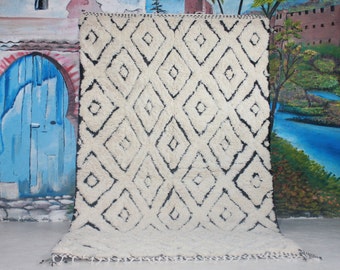 May include: A large, rectangular, cream-colored rug with a repeating pattern of black diamond shapes. The rug has a thick, textured pile and a fringe along the bottom edge. The design is simple yet striking, suitable for a living room or bedroom.