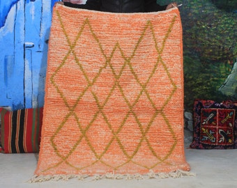 May include: An orange rug with a diamond pattern in a lighter shade of orange and olive green. The rug has a textured appearance and a fringe along the bottom edge. The rug is held up against a blue and green background.