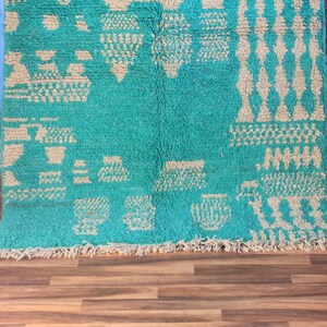 May include: A turquoise and cream-coloured rug with geometric patterns. The rug has a plush texture and a fringe along the bottom edge. The design includes diamond, triangle, and square shapes.