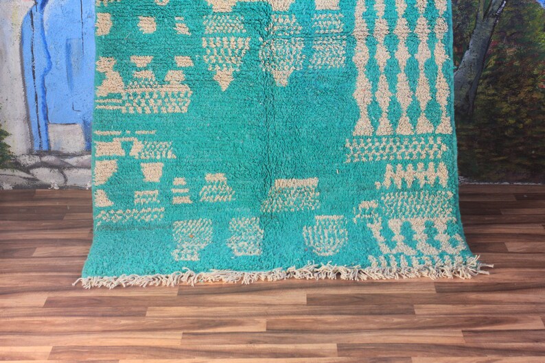 May include: A turquoise and cream-coloured rug with geometric patterns. The rug has a plush texture and a fringe along the bottom edge. The design includes diamond, triangle, and square shapes.