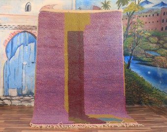 May include: A rectangular, hand-woven rug with a textured surface. The rug features a central vertical design in shades of purple, burgundy, and olive green. The bottom edge has a fringe. The rug is displayed against a painted backdrop.