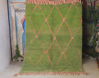 May include: A rectangular green rug featuring a repeating diamond pattern outlined in thin pink lines. The rug has a textured, shaggy pile surface and is finished with short, light-colored fringe along the top and bottom edges.