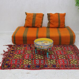 May include: An orange and green striped floor cushion with two matching square pillows. A small, round, yellow table with a colorful design sits on a red patterned rug. A matching orange bag hangs on the wall.