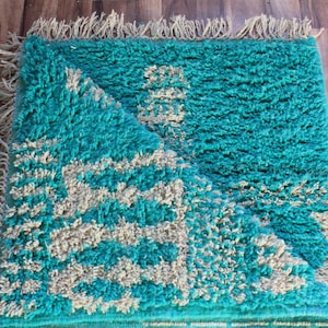 May include: A turquoise rug with a geometric pattern in off-white. The rug has a shaggy texture and fringe along the edges. The rug is folded over, revealing the underside. The rug is rectangular.