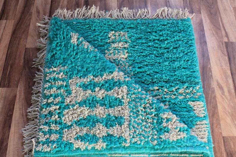 May include: A turquoise rug with a geometric pattern in off-white. The rug has a shaggy texture and fringe along the edges. The rug is folded over, revealing the underside. The rug is rectangular.