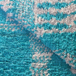 May include: A thick, shaggy rug featuring a high-pile texture with a repeating pattern of vibrant turquoise and soft white yarn. The rug is folded over, revealing the dense, tufted construction and the consistent colour distribution throughout.