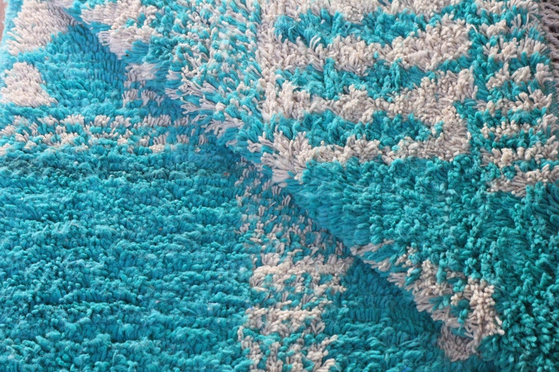 May include: A thick, shaggy rug featuring a high-pile texture with a repeating pattern of vibrant turquoise and soft white yarn. The rug is folded over, revealing the dense, tufted construction and the consistent colour distribution throughout.