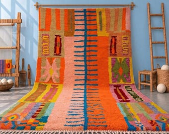 May include: A large rectangular area rug featuring a vibrant, multi-coloured geometric pattern with a central vertical blue spine. The design includes blocks of orange, pink, yellow, and green, finished with a knotted fringe along the bottom edge.