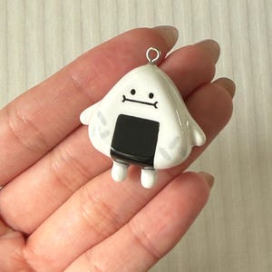 May include: A white, triangular-shaped charm resembling an onigiri rice ball with a black seaweed strip, small arms, and a simple black line face. The charm features a silver-toned metal jump ring at the top for attachment to jewelry or accessories.