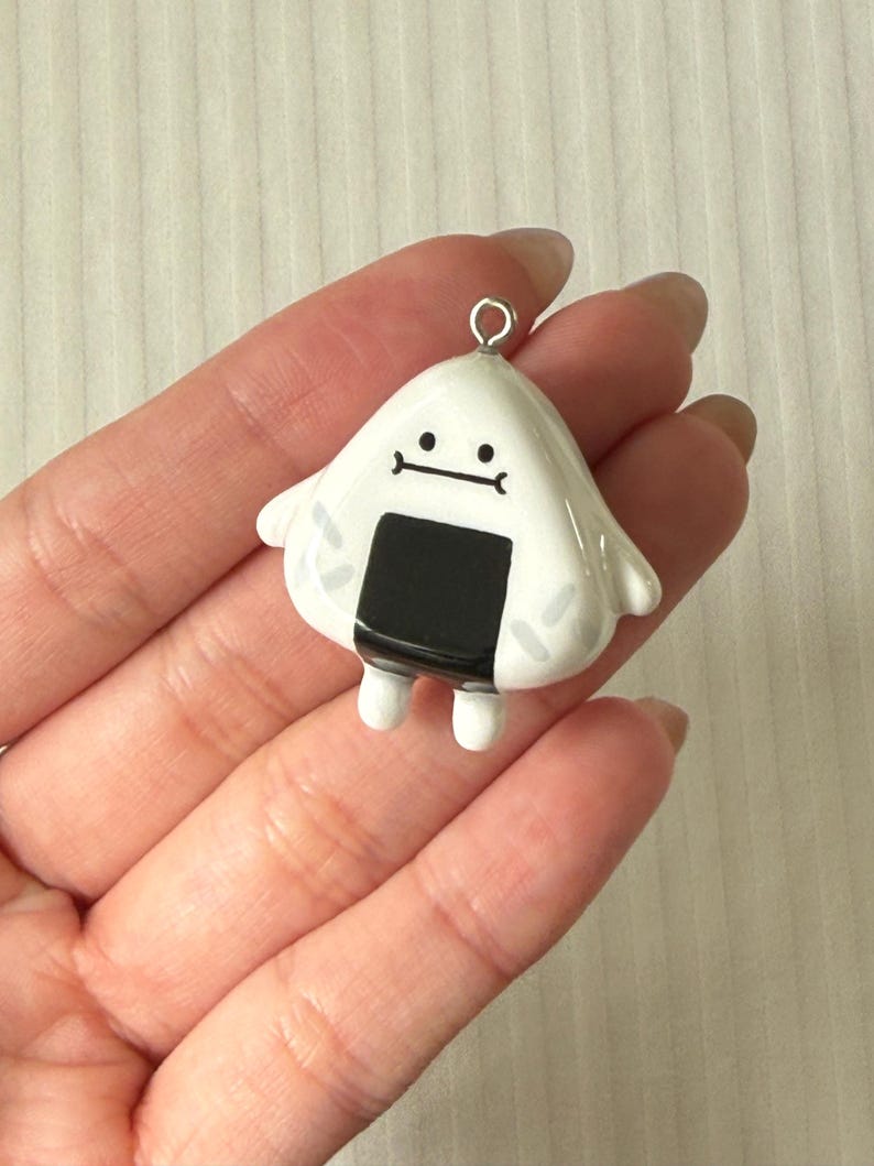May include: A white, triangular-shaped charm resembling an onigiri rice ball with a black seaweed strip, small arms, and a simple black line face. The charm features a silver-toned metal jump ring at the top for attachment to jewelry or accessories.