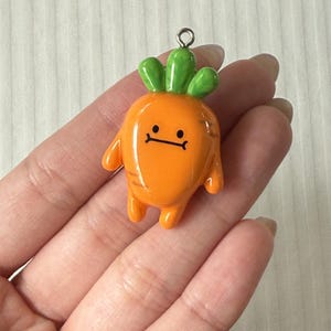 May include: A glossy orange carrot charm with a smooth finish, featuring a green leafy top, two small arms, two small legs, and a simple black line face with two dots for eyes and a straight line for a mouth, topped with a silver metal jump ring.