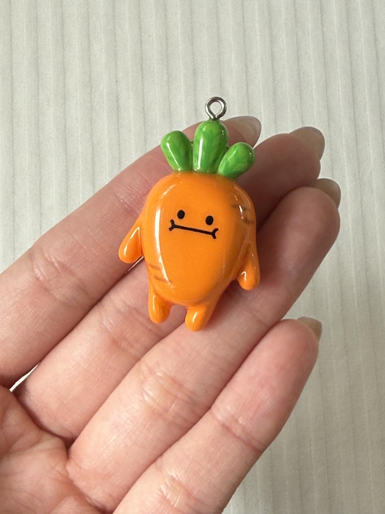 May include: A glossy orange carrot charm with a smooth finish, featuring a green leafy top, two small arms, two small legs, and a simple black line face with two dots for eyes and a straight line for a mouth, topped with a silver metal jump ring.