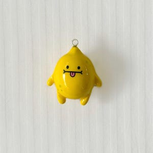 May include: A glossy yellow lemon-shaped charm featuring a small silver metal loop at the top. The charm has a cartoon face with two black dot eyes, a straight black line for a mouth, and a small pink tongue sticking out, with tiny green spots.