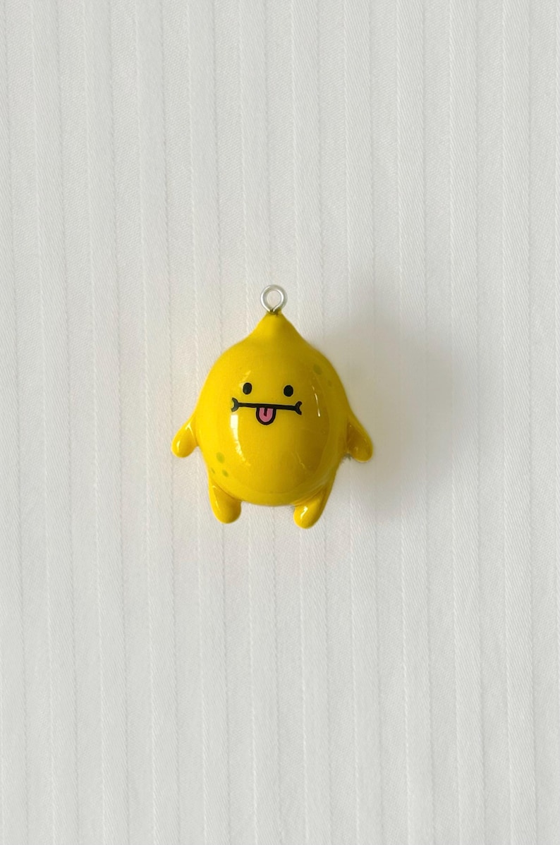 May include: A glossy yellow lemon-shaped charm featuring a small silver metal loop at the top. The charm has a cartoon face with two black dot eyes, a straight black line for a mouth, and a small pink tongue sticking out, with tiny green spots.