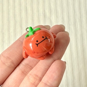 May include: A small, glossy, orange-red tomato charm with a green stem top and a simple black line drawing of a face featuring two dots for eyes and a straight horizontal line for a mouth, including a small silver metal loop at the top.