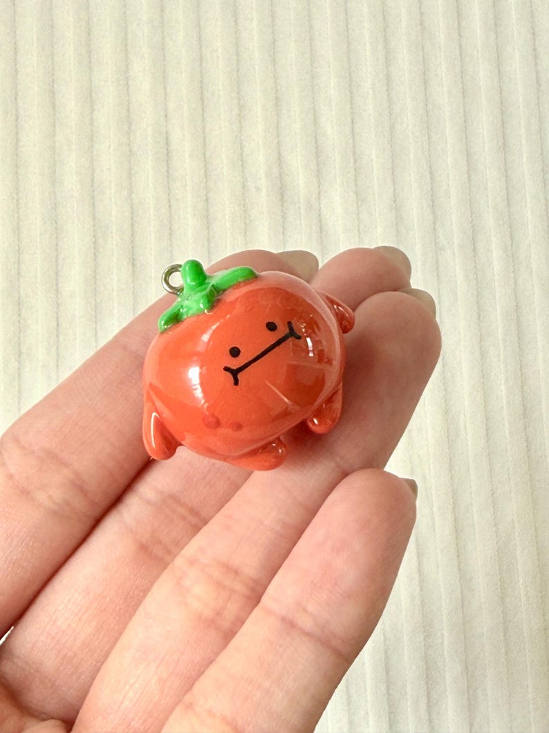 May include: A small, glossy, orange-red tomato charm with a green stem top and a simple black line drawing of a face featuring two dots for eyes and a straight horizontal line for a mouth, including a small silver metal loop at the top.