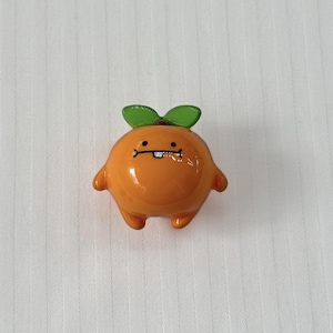 May include: A small, round, orange figurine shaped like a citrus fruit with a smooth, glossy finish. It features two green leaves on top, two small arms, two short legs, and a printed face with two black eyes and a single white tooth.