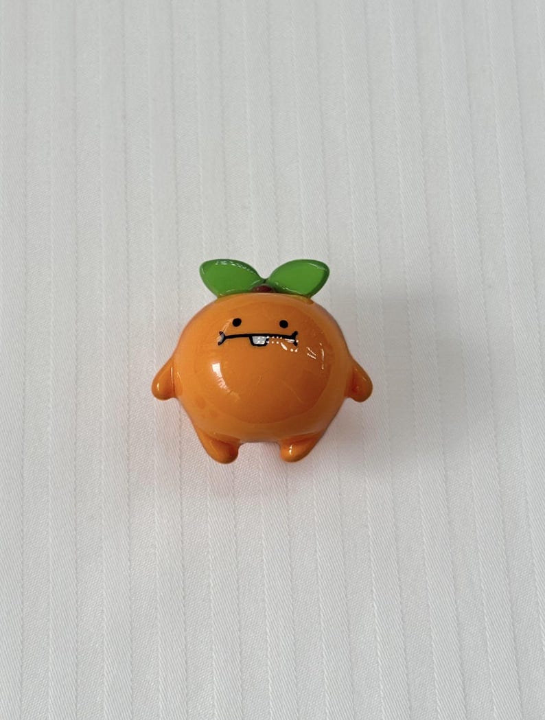 May include: A small, round, orange figurine shaped like a citrus fruit with a smooth, glossy finish. It features two green leaves on top, two small arms, two short legs, and a printed face with two black eyes and a single white tooth.