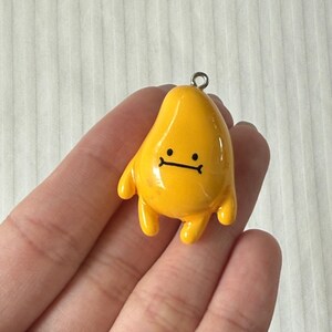 May include: A glossy yellow charm shaped like a teardrop or abstract character with a simple black line drawing of a face featuring two dots for eyes and a straight line for a mouth. The charm includes a small silver metal loop at the top.