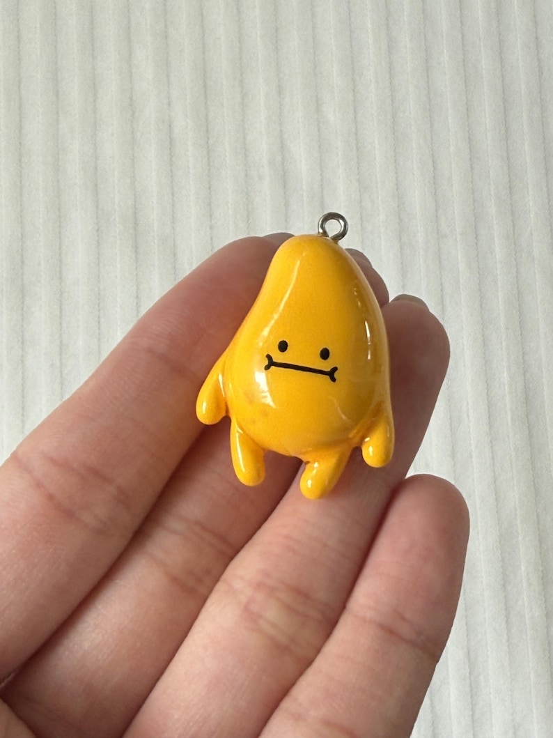 May include: A glossy yellow charm shaped like a teardrop or abstract character with a simple black line drawing of a face featuring two dots for eyes and a straight line for a mouth. The charm includes a small silver metal loop at the top.
