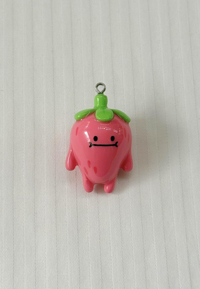 May include: A glossy pink strawberry-shaped charm with a smooth finish, featuring a green stem top, two small arms, two legs, and a simple black line face with two dots for eyes. A small silver metal jump ring is attached to the top stem.