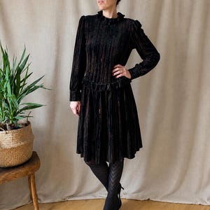 Può includere: Un abito in velluto nero con maniche lunghe, colletto a balze e un dettaglio a balze in vita. Il tessuto presenta una trama a righe verticali con sottili riflessi color rame. L'abito ha una gonna al ginocchio e polsini aderenti.