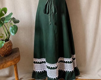 May include: A white ribbed short-sleeve top with a button-down front paired with a floor-length dark green skirt. The skirt features a matching green polka-dot tie belt and two horizontal bands of intricate white lace trim near the hemline.