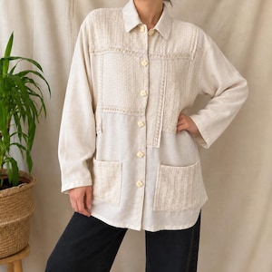 Puede incluir: Una camisa de manga larga en color blanco roto con cuello, dos bolsillos delanteros de parche y una mezcla de paneles de tela texturizada en el pecho y los bolsillos. La camisa se cierra con botones cuadrados de color claro.