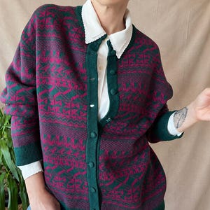 Könnte beinhalten: Eine langärmelige Strickjacke mit Knopfleiste in einem dunkelgrünen und magentafarbenen geometrischen Muster, getragen über einem weißen Hemd mit gewelltem Kragen und einer blauen Jeans. Die Strickjacke hat gerippte Bündchen und passende Stoffknöpfe.