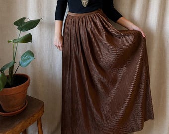 May include: A long, brown, pleated skirt with a metallic sheen, paired with a black, long-sleeved, ribbed knit top and a gold-toned pendant necklace featuring a feline head design. The skirt reaches the floor and has a gathered waistband.