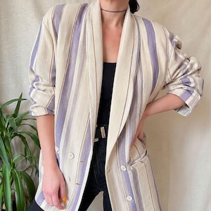 May include: A long-sleeve blazer featuring a vertical striped pattern in shades of cream, light purple, and thin brown lines. The garment has a shawl collar, a double-breasted front closure with four visible buttons, and two side pockets.