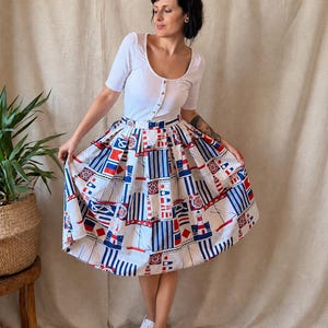 May include: A white ribbed short-sleeve top with a button-down front paired with a pleated midi skirt featuring a nautical print. The skirt pattern includes red, white, and blue illustrations of sailboats, lighthouses, and maritime signal flags.
