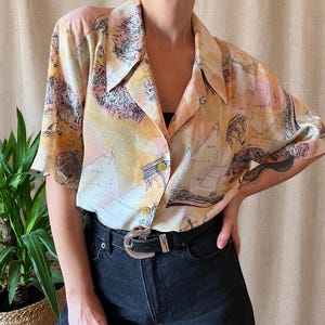 May include: A short-sleeved button-down shirt featuring a light yellow, peach, and white watercolor-style print of sailboats and coastal scenery. The shirt has a wide collar, two visible buttons, and is paired with black denim jeans and a black belt.