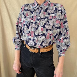 Könnte beinhalten: Ein langärmliges Hemd mit geometrischem Muster in Marineblau, Rosa, Grau und Weiß. Das Hemd hat einen Kragen und zwei Brusttaschen. Das Model trägt eine schwarze Jeans und einen beigefarbenen Ledergürtel mit silberner Schnalle.